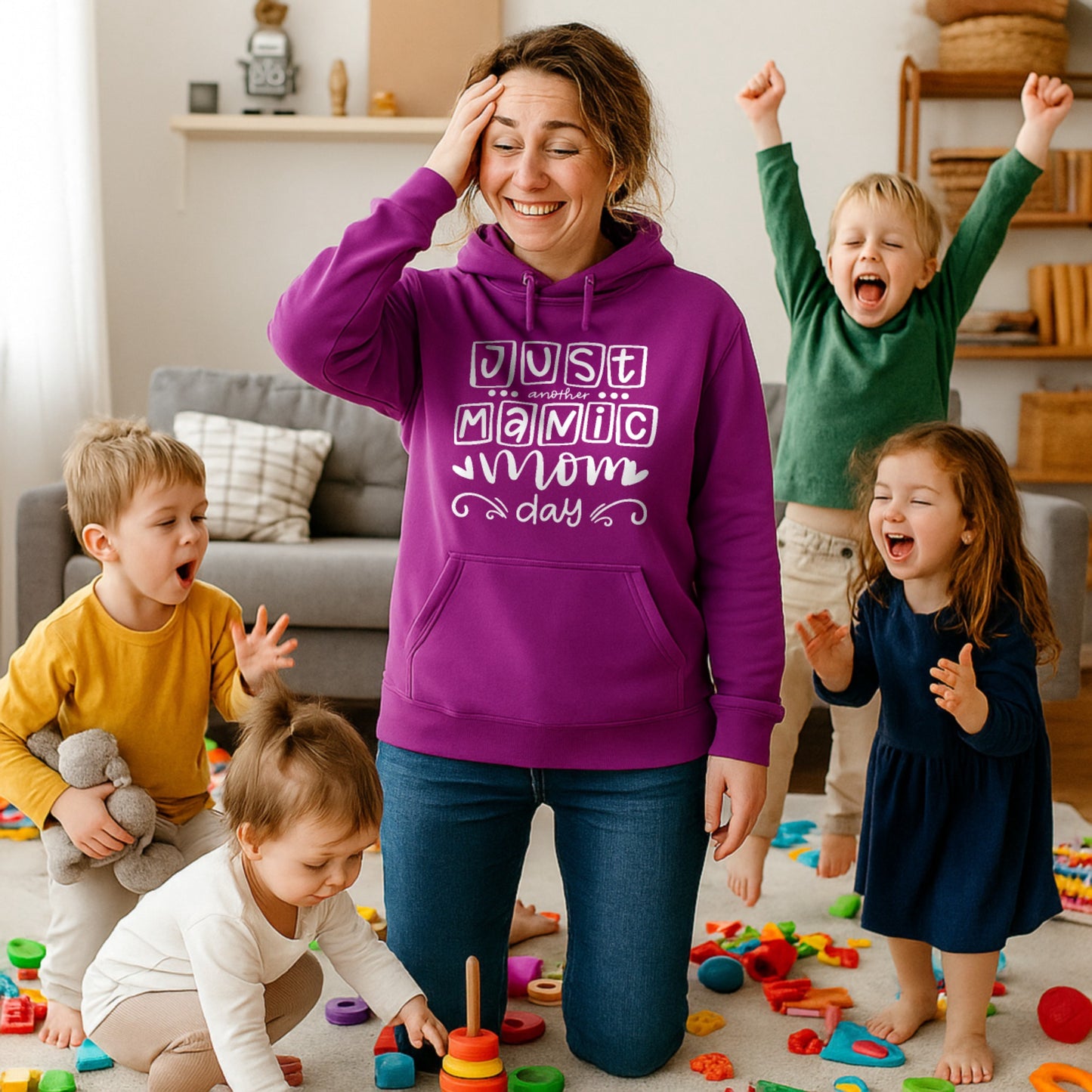 Smiling mum wearing a pinky purple "Just Another Manic Mom Day" hoodie while surrounded by energetic children and colourful toys. Funny mom life hoodie celebrating the chaos of motherhood, perfect as a mom gift or Mother's Day gift.