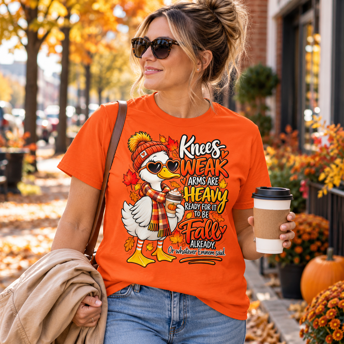 Woman wearing an orange Knees Weak Arms Are Heavy funny autumn goose T-shirt while carrying coffee on a colourful autumn street.