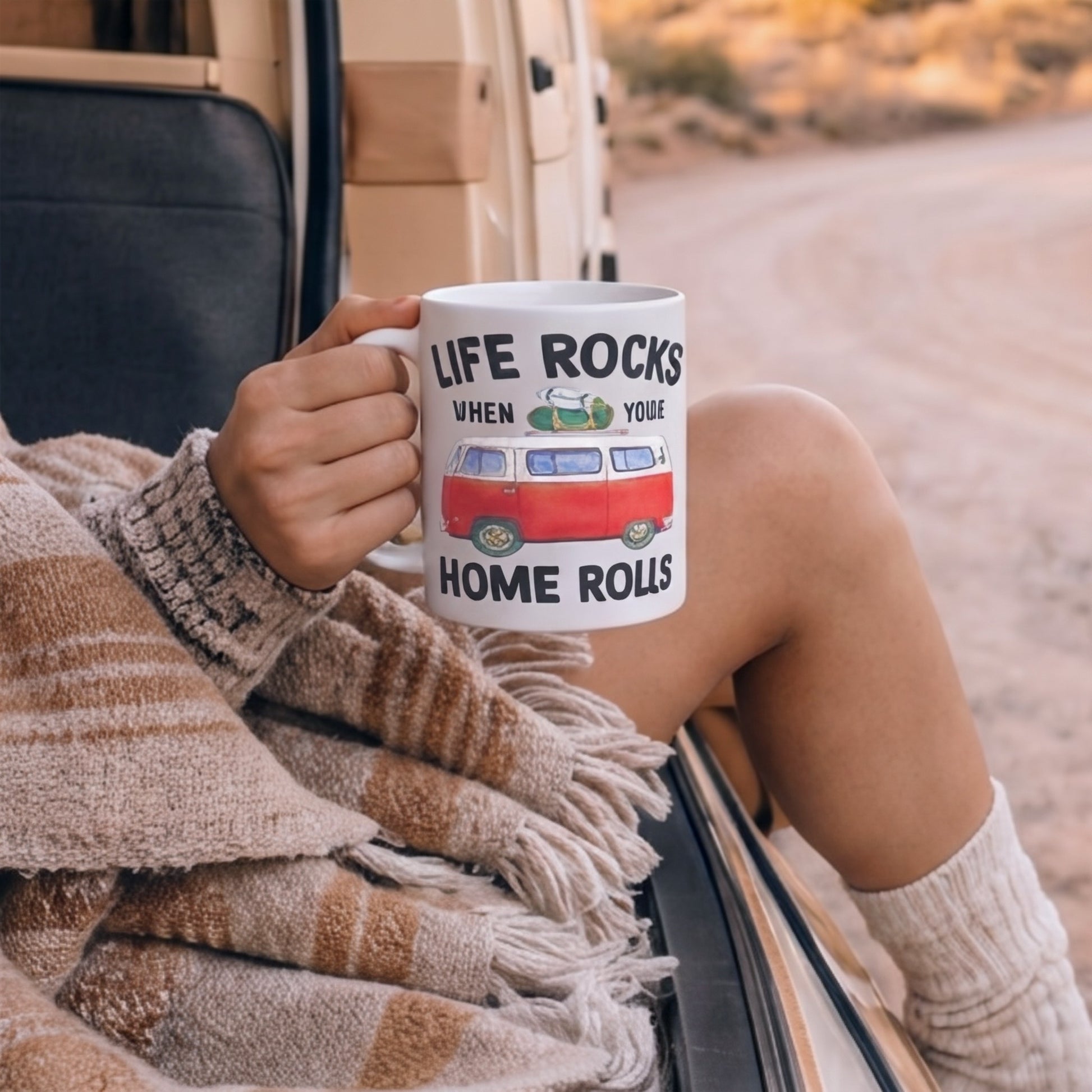 Lifestyle mockup of the Life Rocks When Your Home Rolls camper van mug being held outside a campervan, featuring a vintage red camper van design during a cosy van life coffee break on a scenic road trip.