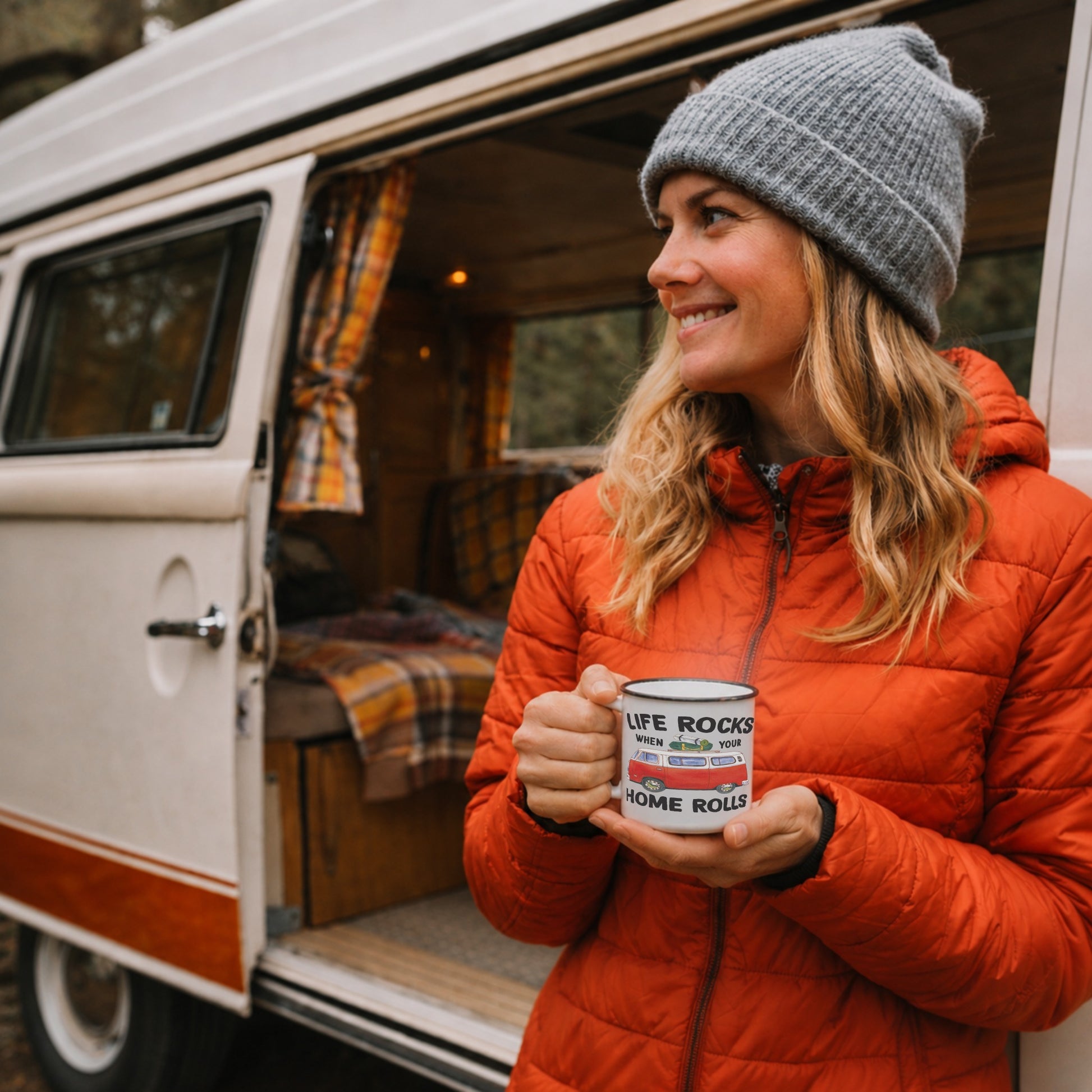 Lifestyle mockup of the Life Rocks When Your Home Rolls outdoor enamel mug held by a woman outside a vintage campervan, featuring a red camper van design during a cosy camping and van life adventure.