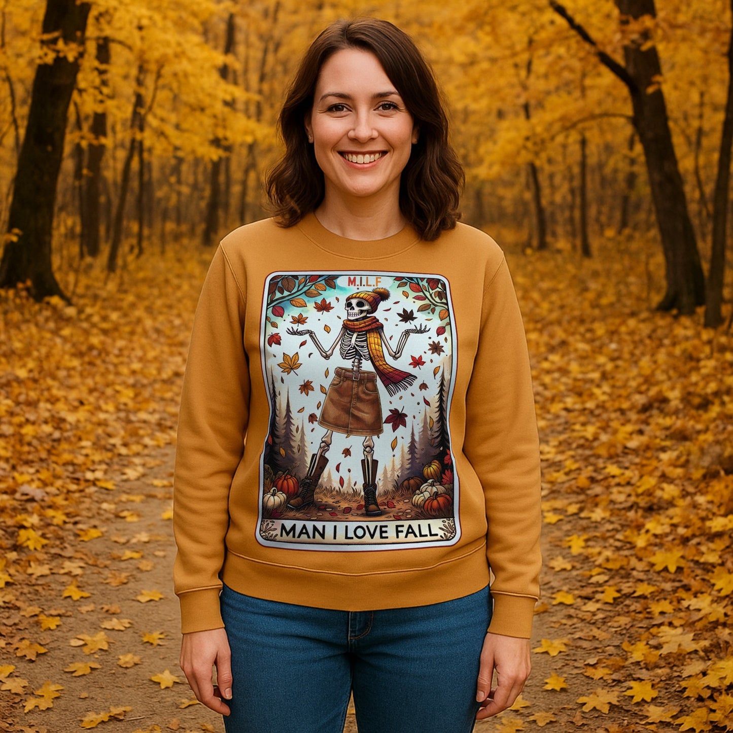 Woman wearing a mustard-coloured MILF Man I Love Fall sweatshirt featuring a skeleton, pumpkins, and autumn leaves while standing on a forest path surrounded by vibrant fall foliage.