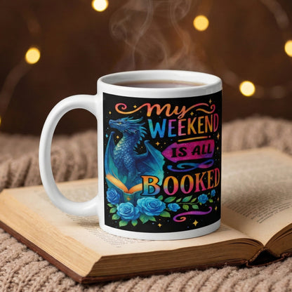 My Weekend Is All Booked book lover coffee mug featuring a blue fantasy dragon and enchanted book design resting on an open book with hot coffee in a cosy reading nook.