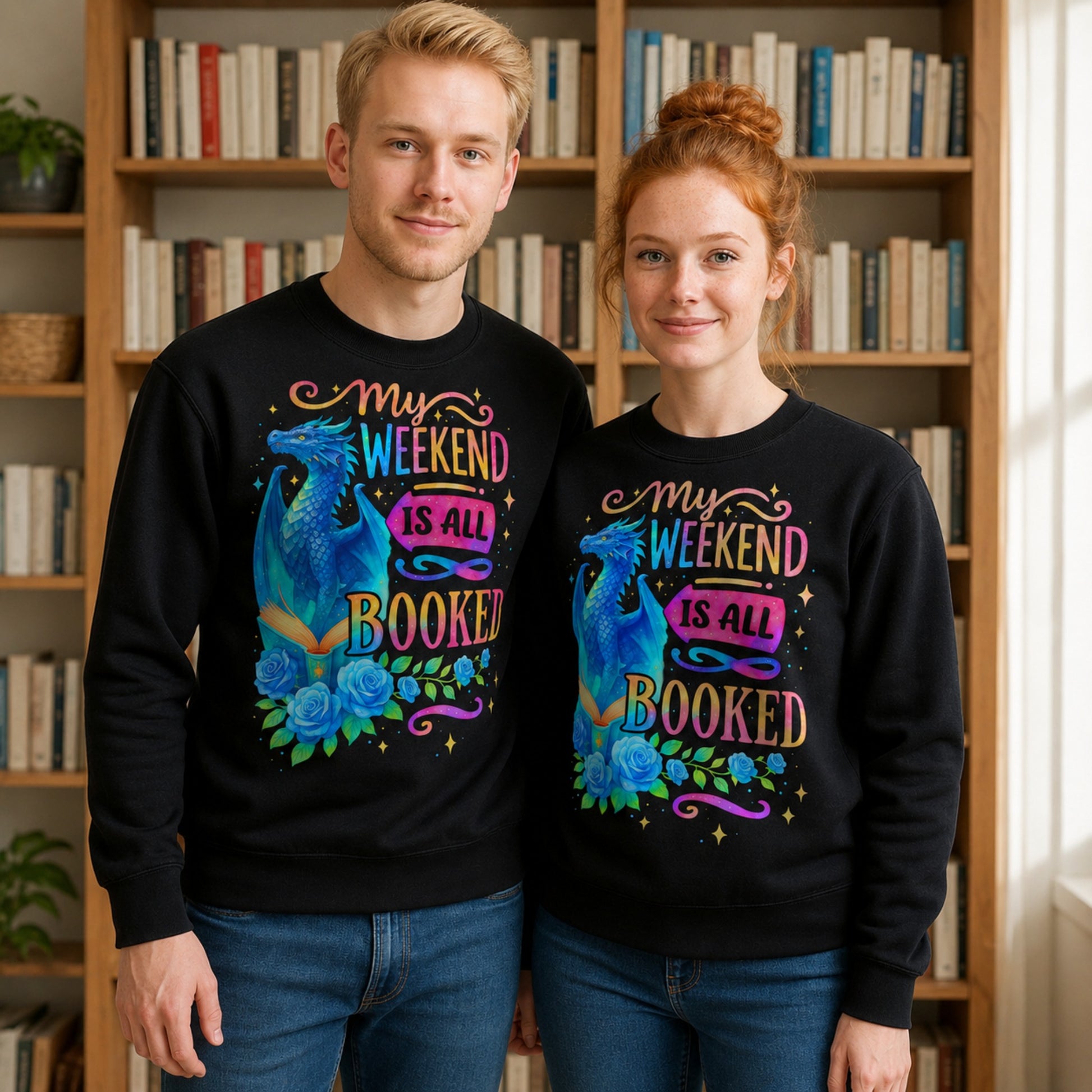 Male and female models wearing matching black My Weekend Is All Booked sweatshirts featuring a colourful blue dragon, open book, roses, and fantasy-inspired book lover design while standing in front of bookshelves.