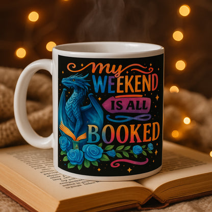 Mug with dragon design and 'My weekend is all booked' text on an open book with warm lighting.