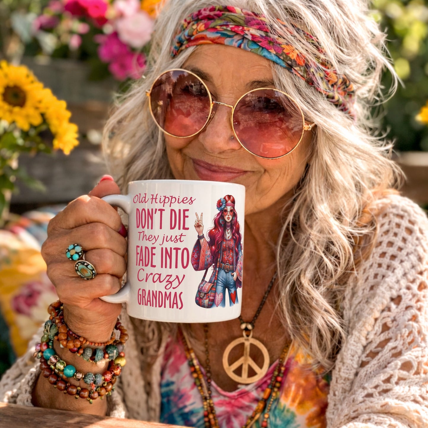 Lifestyle mockup of the Old Hippies Don't Die They Just Fade Into Crazy Grandmas funny grandma mug being held by a smiling retro hippie woman wearing round sunglasses, a colourful headband, and peace necklace in a vibrant flower-filled garden.