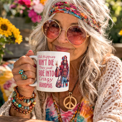 Lifestyle mockup of the Old Hippies Don't Die They Just Fade Into Crazy Grandmas funny grandma mug being held by a smiling retro hippie woman wearing round sunglasses, a colourful headband, and peace necklace in a vibrant flower-filled garden.