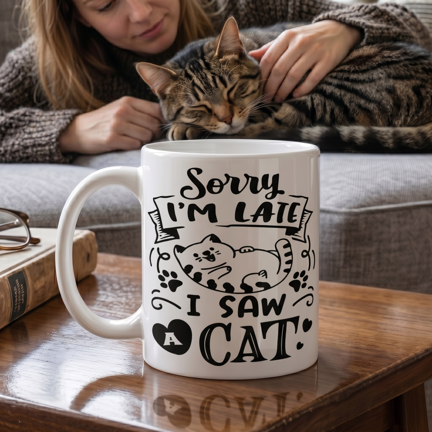 White coffee mug featuring the phrase "Sorry I'm Late, I Saw A Cat" on a wooden table while a woman cuddles a sleeping tabby cat in the background.