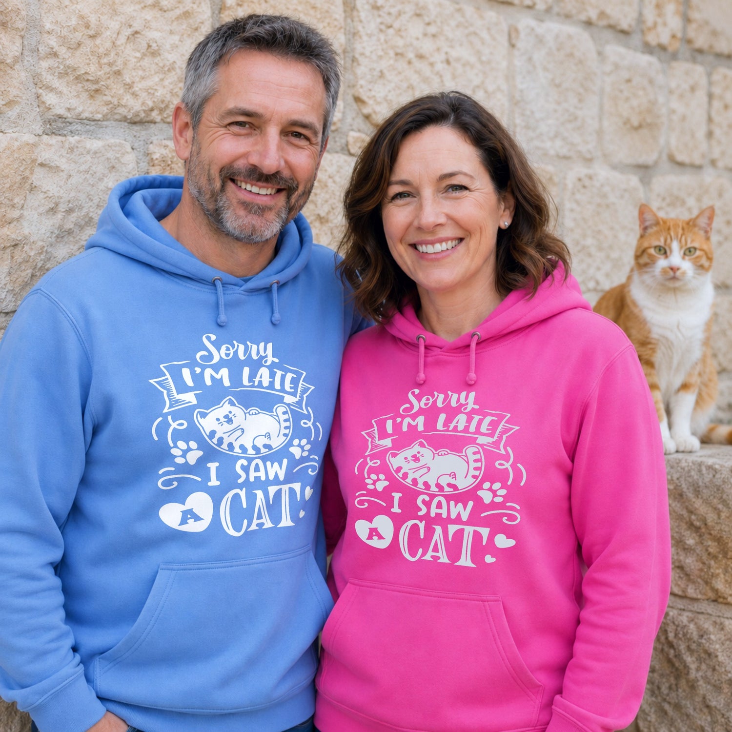 Two adults wearing matching blue and pink hoodies featuring the slogan "Sorry I'm Late, I Saw a Cat" stand smiling beside a ginger cat against a stone wall, showcasing a funny cat lover hoodie design.