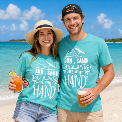 Smiling couple wearing matching Sun Sand And A Drink In My Hand t-shirts on a tropical beach while holding drinks, perfect for summer vacations, cruises, and beach lovers.