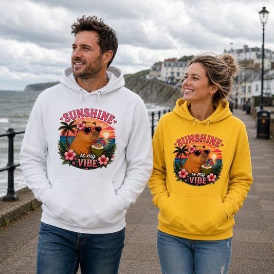 Man wearing a white Sunshine Is My Vibe Capybara Hoodie and woman wearing a yellow Sunshine Is My Vibe Capybara Hoodie walking along a British seafront promenade with coastal views on a cloudy day.