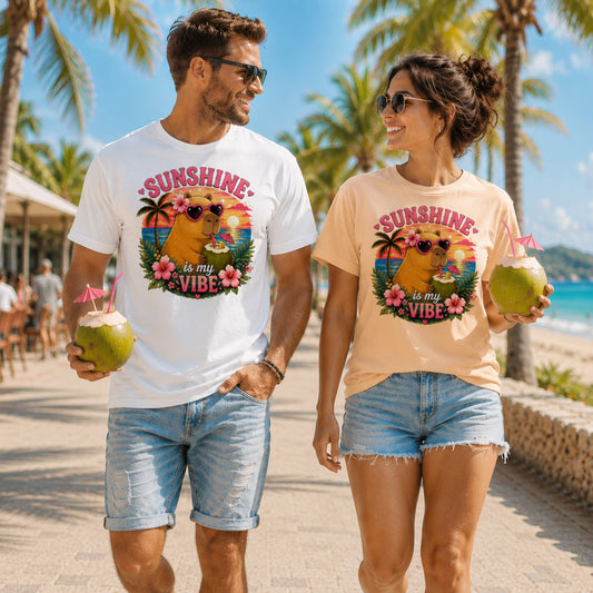 Couple wearing Sunshine Is My Vibe Capybara T-Shirts on a tropical beach promenade, featuring colourful capybara graphic tees with coconut drinks, palm trees and sunny holiday vibes.