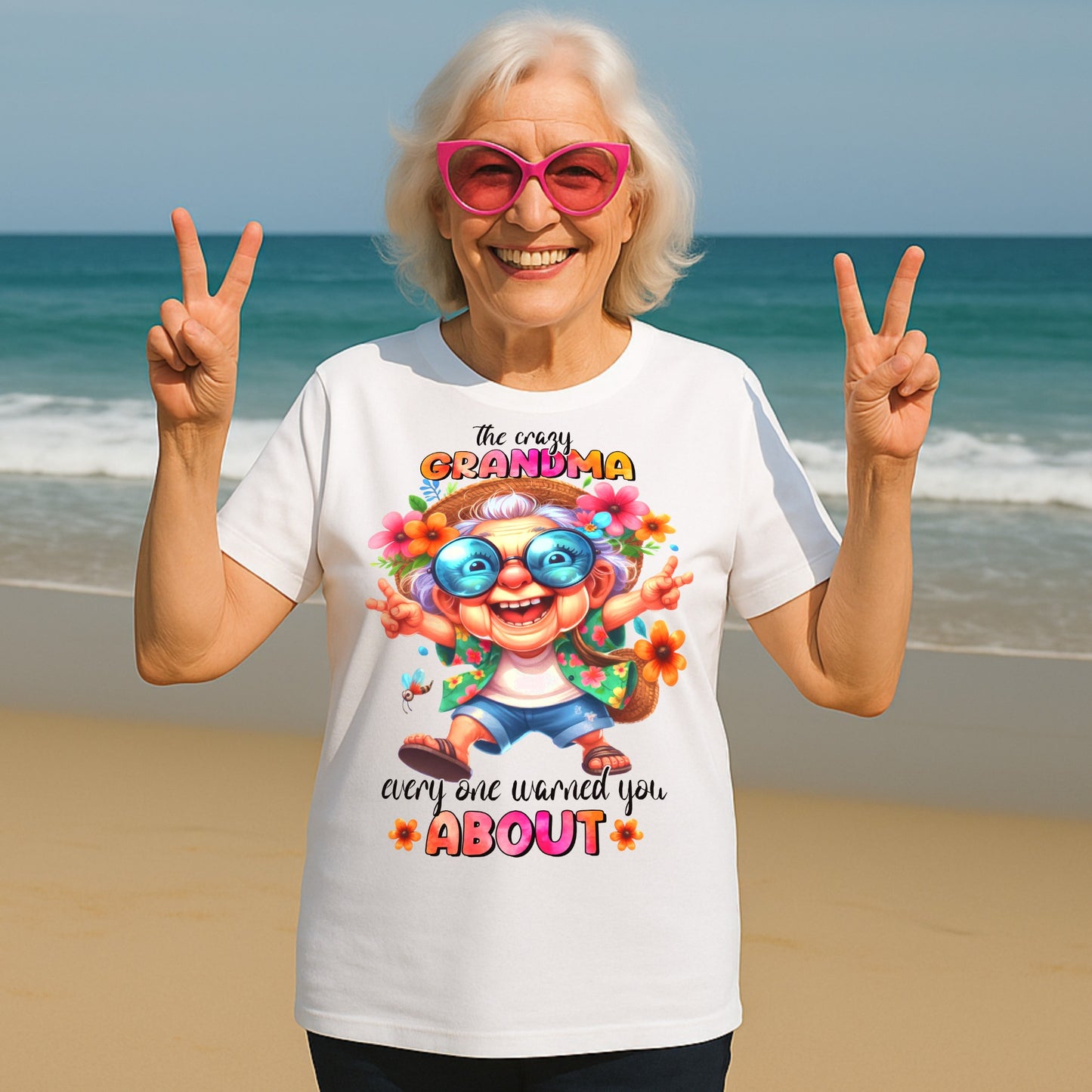 Smiling senior woman wearing a funny grandma shirt with "The Crazy Grandma Everyone Warned You About" design, posing on a beach in pink sunglasses and making peace signs.