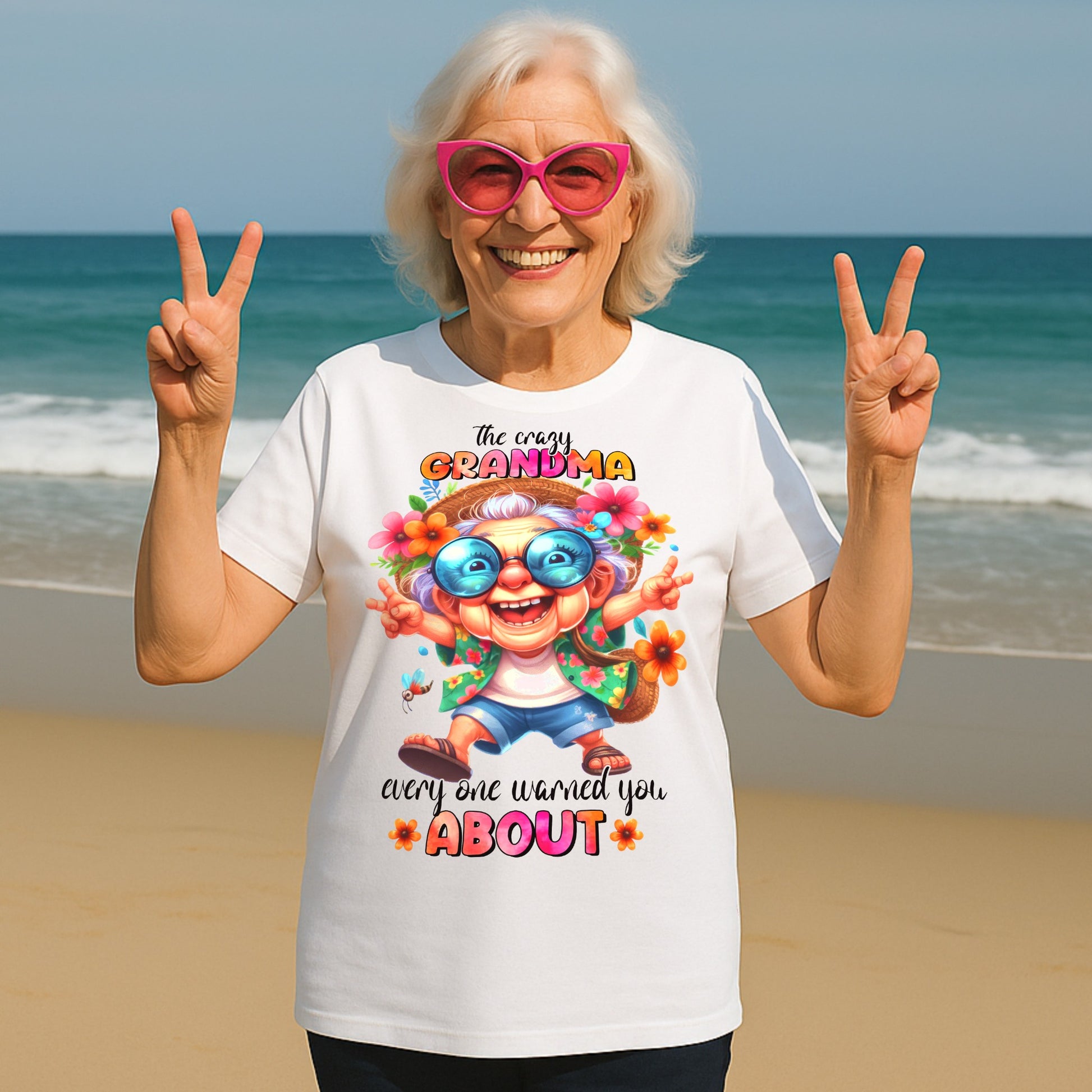 Smiling senior woman wearing a funny grandma shirt with "The Crazy Grandma Everyone Warned You About" design, posing on a beach in pink sunglasses and making peace signs.