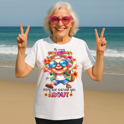 Smiling senior woman wearing a funny grandma shirt with "The Crazy Grandma Everyone Warned You About" design, posing on a beach in pink sunglasses and making peace signs.