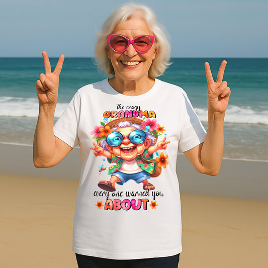 Smiling senior woman wearing a funny grandma shirt with "The Crazy Grandma Everyone Warned You About" design, posing on a beach in pink sunglasses and making peace signs.