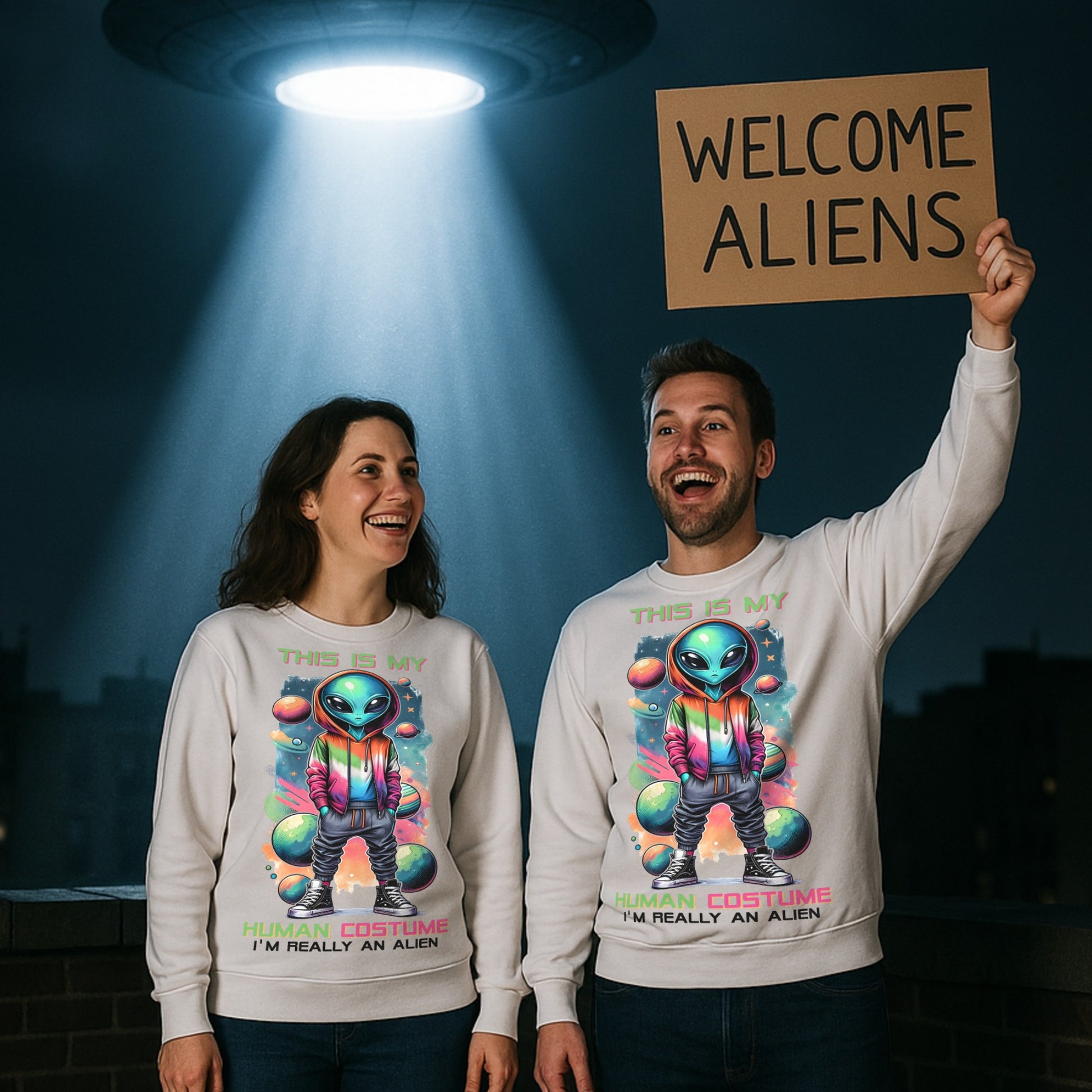 Two people wearing sweatshirts with alien graphics, one holding a 'Welcome Aliens' sign. They are wearing a white sweatshirt with colourful galaxy design the text reads this is my human costume I'm really an alien. 
