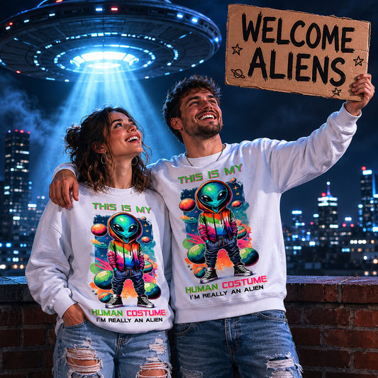 Man and woman wearing matching "This Is My Human Costume I'm Really An Alien" sweatshirts beneath a glowing UFO while holding a Welcome Aliens sign in a city night scene.