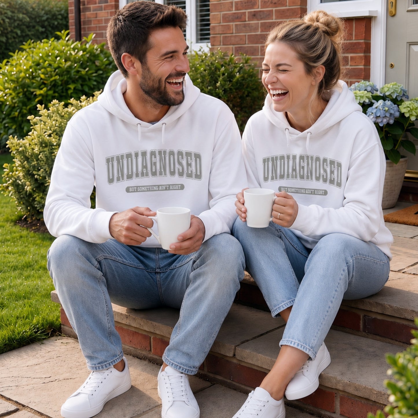 Couple wearing matching white "Undiagnosed But Something Ain't Right" hoodies sitting on front porch steps holding plain white coffee mugs and laughing together in a realistic lifestyle mockup.